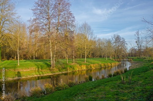 A river is winding in the middle of a meadow. Trees are naked. The sun is setting, illuminating the landscape with a golden light. White clouds are scattered in the blue sky.