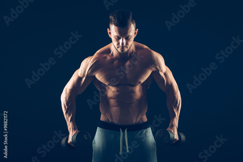 Closeup of a muscular young man lifting dumbbells weights on dark background