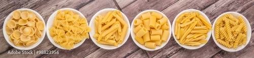 Obraz  z motywem Various dried pasta variety and shapes in white bowl over wooden background
