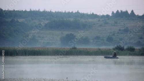 Fisherman on an inflatable boat in the early morning fishing on a lake. Mist rises over the water.