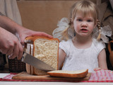 Ребенок смотрит как нарезают хлеб. На столе свежеиспеченный хлеб. Мужские руки режут хлеб большим ножом. Огромная буханка белого хлеба и маленькая девочка с двумя косичками.