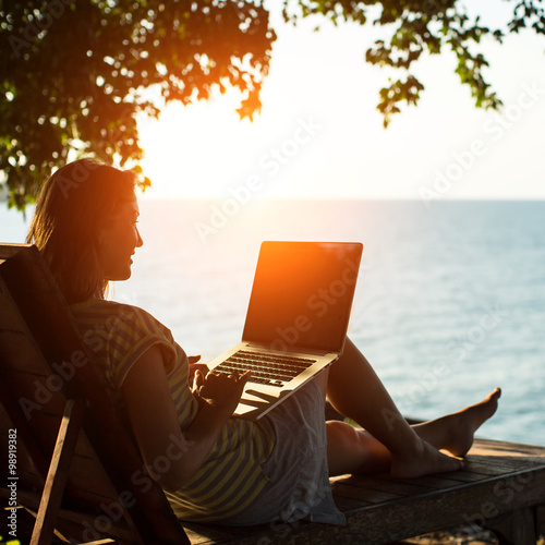 Fotografie Silhouette girl c laptop sitting on a lounger near the sea in the sunset