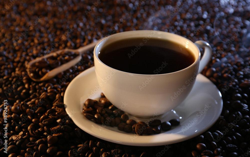 Obraz premium Steaming hot black coffee in white ceramic cup with saucer on roasted coffee beans and wooden scoop