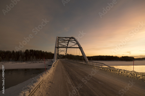 A bridge on a river partly frozen with a sunset in the background