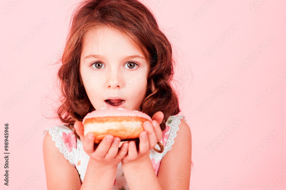Cute little girl eating donut over pink. Looking at camera. Childhood. 