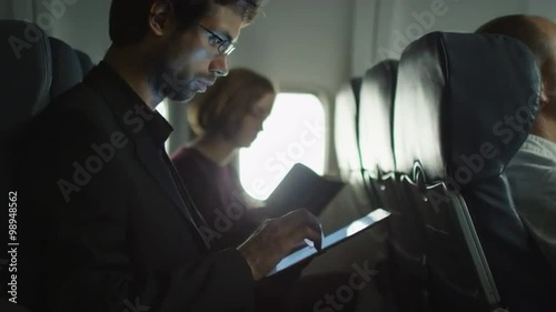 Young man is using a tablet on an airplane and a woman is reading in the background next to a window.