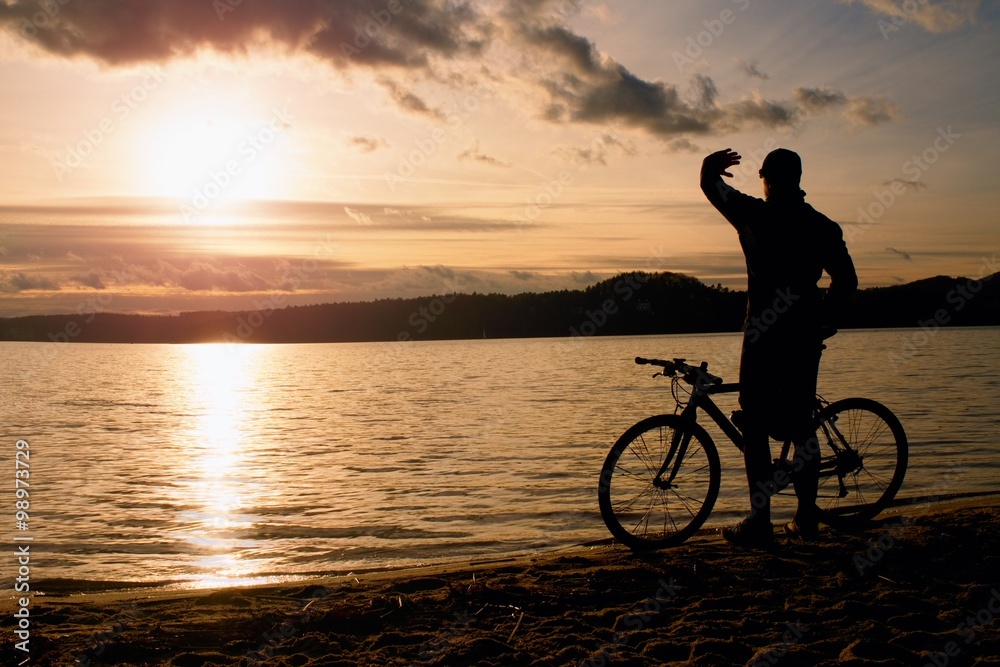 Obraz premium Young man cyclist silhouette on blue sky and sunset above the beach. Biker atthe end of season at lake.