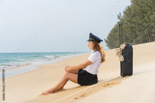 Travel concept. Attractive flight attendant with suitcase, hat and shoes on the beach.