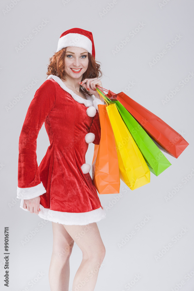 Christmas and New Year Concept and Ideas. Sexy Looking Young Caucasian Red Haired Female in Santa Hat Posing with Shopping Bags Against White Background