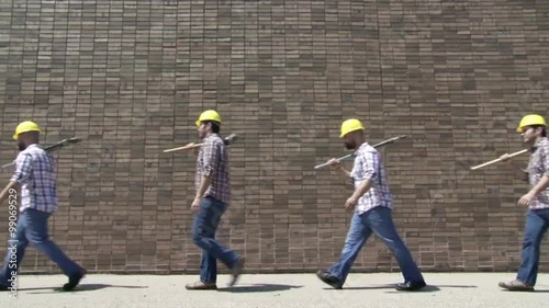 Two contractors with a sledgehammer and an adjustable wrench walk endlessly past the same brick wall.  Locked off wide shot.  Looping clip. photo