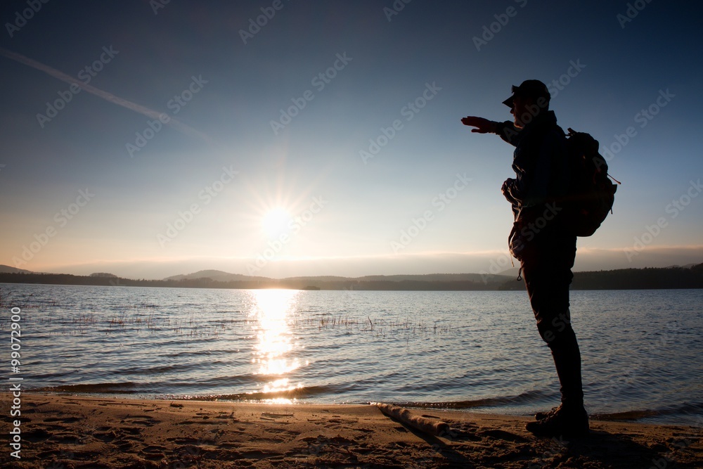 Man with hand in the air. Tall hiker in dark sportswear with sporty backpack stands on beach enjoy sunset at horizon