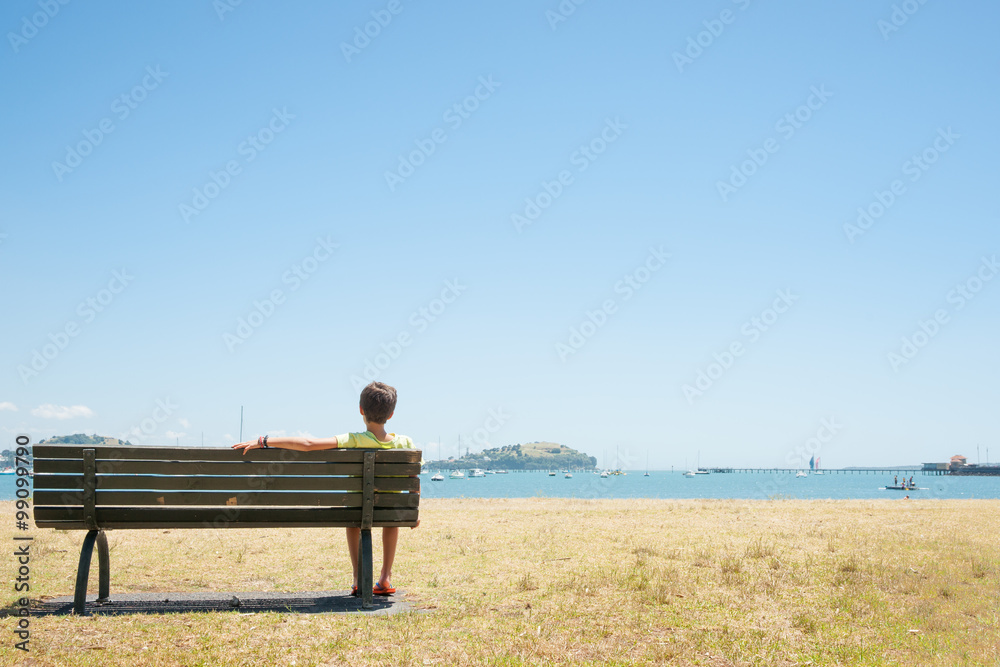 Boy sits on bench seat in the bay. Summer day view across bay, Orakei Auckland.