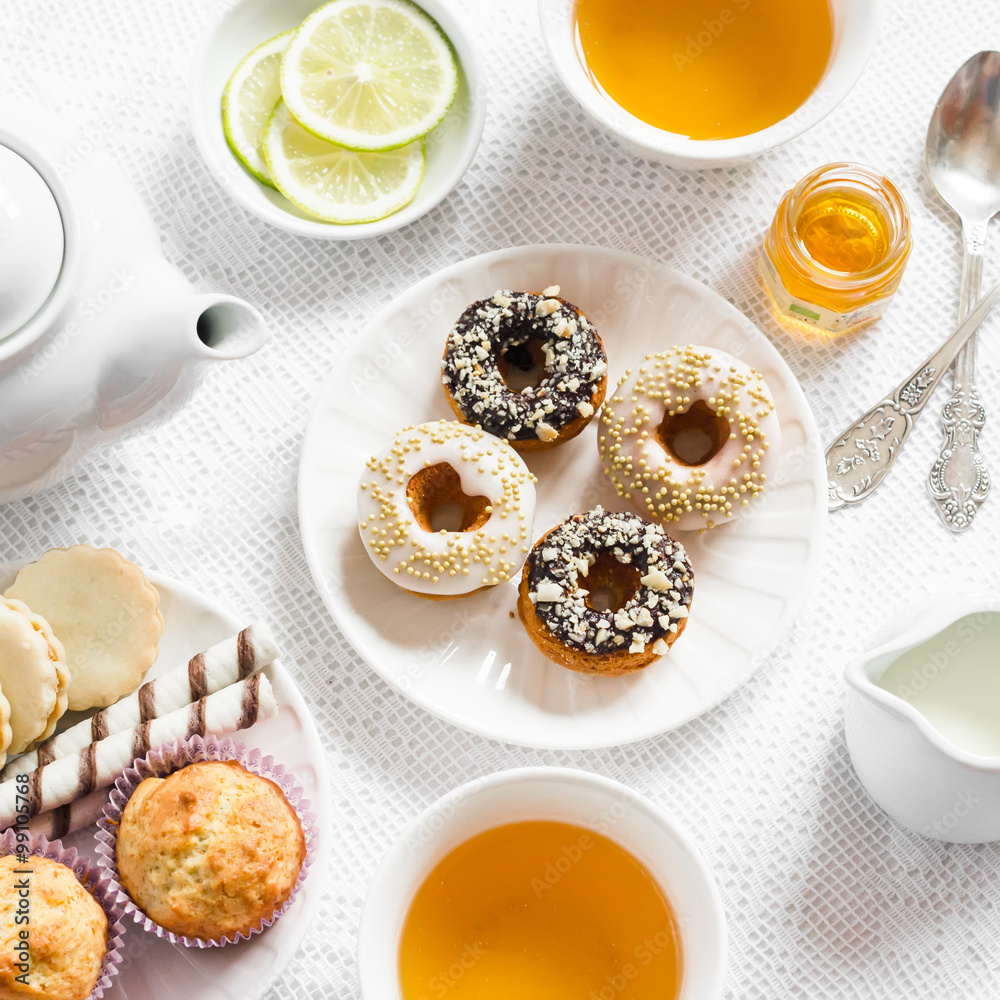 Lemon green tea and sweets - banana muffins, cookies with caramel and nuts, donuts with chocolate and lemon glaze, tea set on white table cloths on a light surface. Tea time. Vintage and rustic style Lemon green tea and sweets - banana muffins, cookies with caramel and nuts, donuts with chocolate and lemon glaze, tea set on white table cloths on a light surface. Tea time. Vintage and rustic style