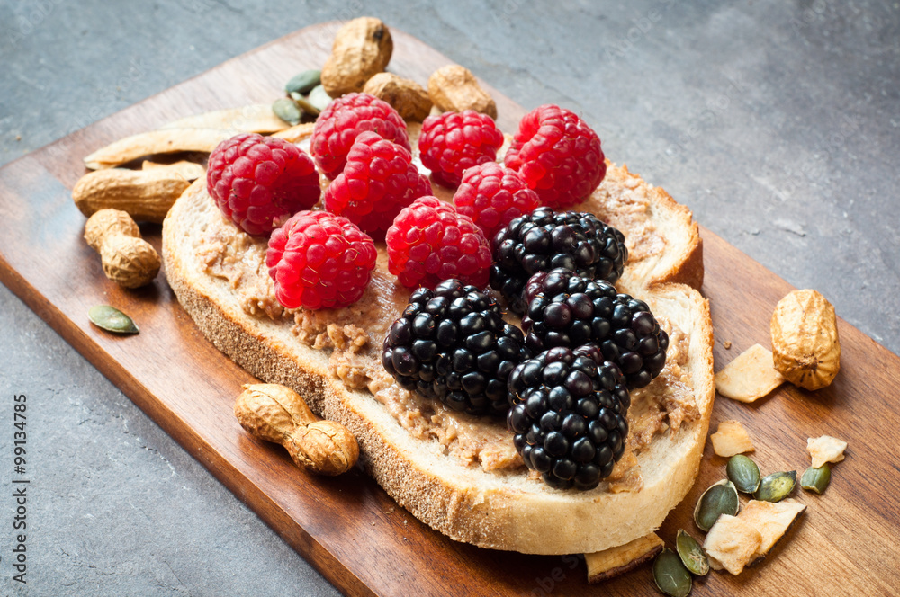 Healthy snack of sliced sourdough bread topped with peanut butter and fresh blackberries and raspberries. Served on a wooden tray on a slate stone table top.