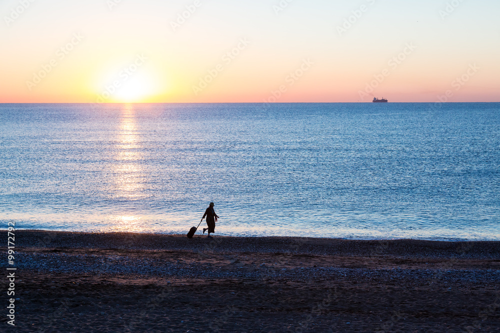 Fototapeta premium Exotic Vacation Silhouette of Female pulling Travel Suitcase along Ocean Beach