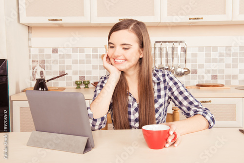 Laughing girl looking into the tablet and holding a cup of tea i