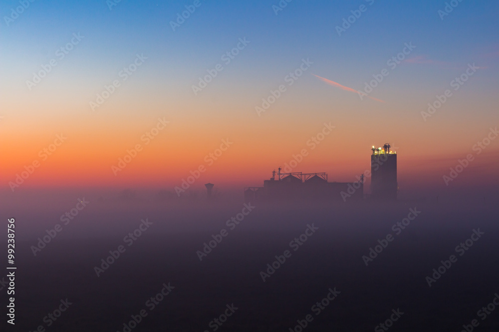 Industrial foggy landscape, silhouette of old factory against the sunset sky and the mist at blue hour at night