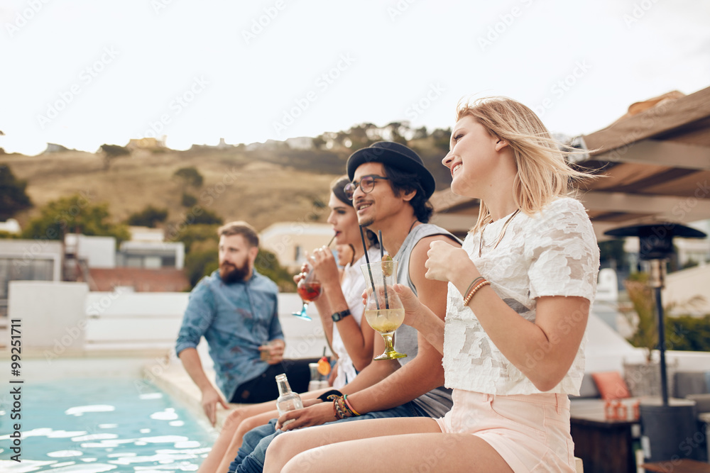 Happy young friends sitting by the pool having drinks