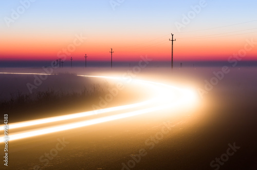 Long Exposure White Car light trails on a road outside at foggy night on blue hour with electrical power lines and pylons disappear over the horizon