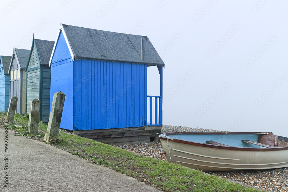 Blue wooden beach huts and old fishing boat by a footpath on British seaside on a foggy day Blue wooden beach huts and old fishing boat by a footpath on British seaside on a foggy day