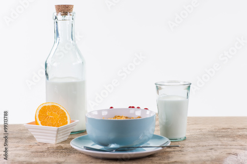 Frühstück mit Müsli in blauer Schale, eine angeschnittene Orange und Milch auf hellem Holztisch