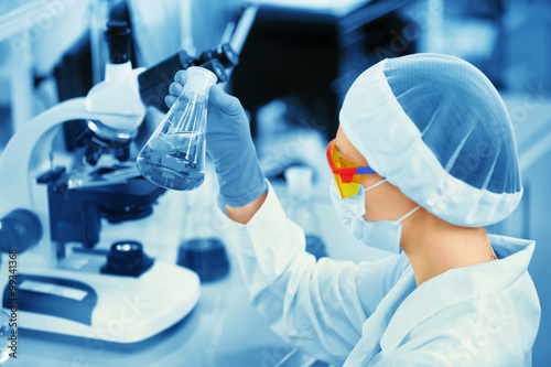 A female medical or scientific researcher or woman doctor looking at a test tube of solution in a laboratory with microscope in background.