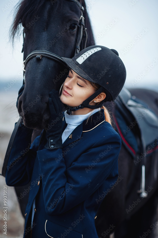 a tall girl with long blond hair in a jockey outfit with a beautiful black horse in an empty snow-covered field in winter a tall girl with long blond hair in a jockey outfit with a beautiful black horse in an empty snow-covered field in winter