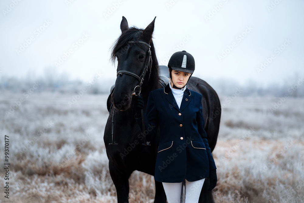 Obraz premium a tall girl with long blond hair in a jockey outfit with a beautiful black horse in an empty snow-covered field in winter