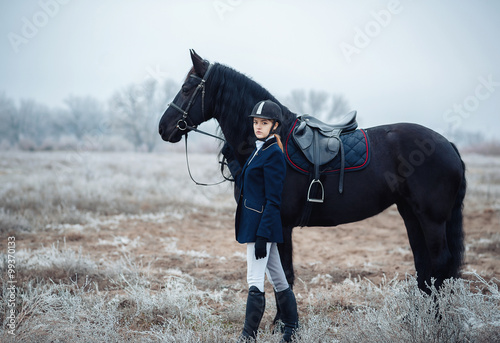 a tall girl with long blond hair in a jockey outfit with a beautiful black horse in an empty snow-covered field in winter