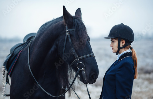 a tall girl with long blond hair in a jockey outfit with a beautiful black horse in an empty snow-covered field in winter
