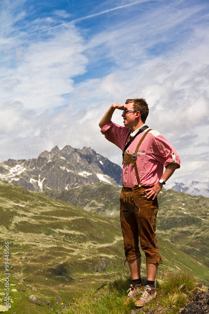 Young explorer in traditional clothing looks out over the valley on top of a mountain in the Swiss Alps on a sunny summer day