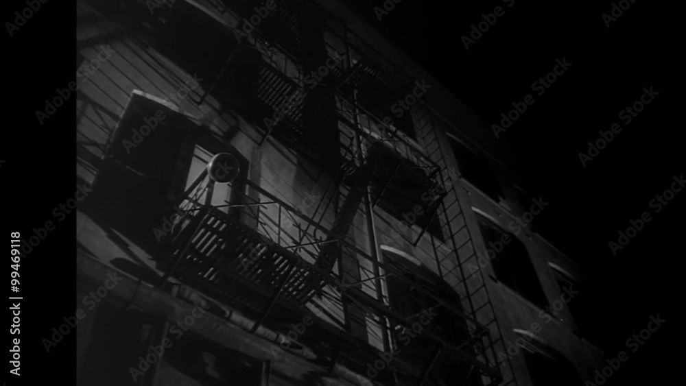 Low angle view of man climbing down fire escape at night, 1940s