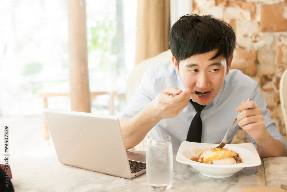 Young Asian man working while eating with his laptop in restaurant (selective focus) Young Asian man working while eating with his laptop in restaurant (selective focus)