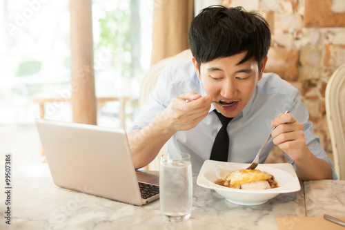Young Asian man working while eating with his laptop in restaurant
