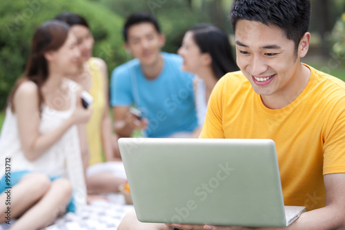 Young man using computer on meadow with friends in background