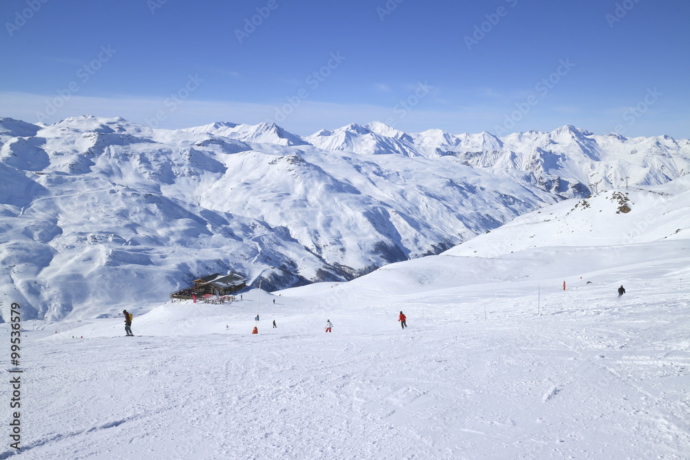 Skiers on ski slopes in high Alps resort, apres ski chalet, with snowy mountain peaks in against blue sky