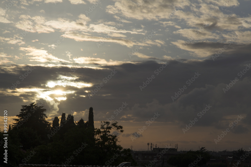 Cityscape in Rome at dusk with sun behind the clouds. Silhouette of trees.