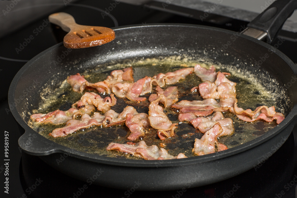 Speck wird in der Pfanne knusprig gebraten. Speck in einer Pfanne mit Fett auf dem Herd. Holzlöffel daneben.
