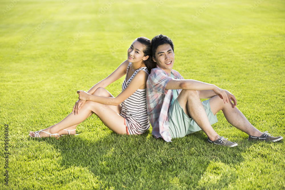Cheerful young couple sitting back to back on grass Cheerful young couple sitting back to back on grass