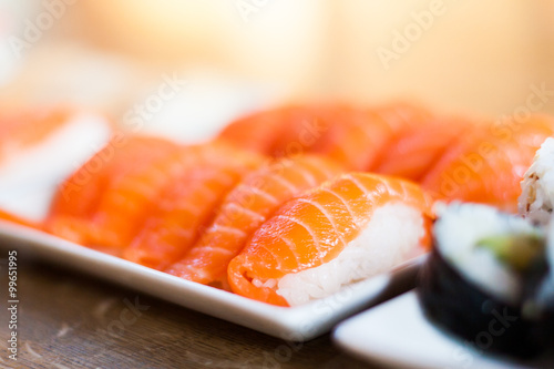 Many pieces of salmon nigiri sushi are places on a nice square plate. You also see some maki rolls in the foreground. 