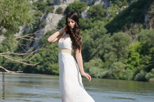 Beautiful young woman wearing a long white dress, standing side by the river