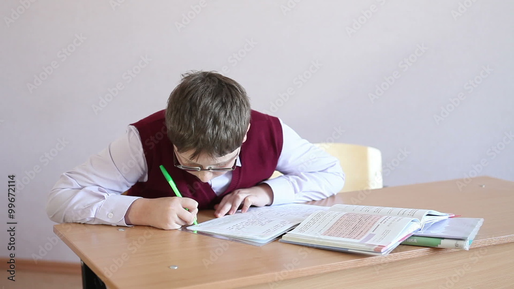 boy with glasses writes in a notebook sitting at a desk boy with glasses writes in a notebook sitting at a desk