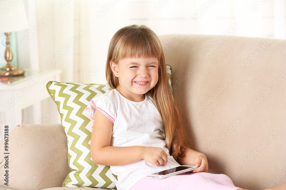 Beautiful little girl with smart phone sitting on sofa, on home interior background Beautiful little girl with smart phone sitting on sofa, on home interior background