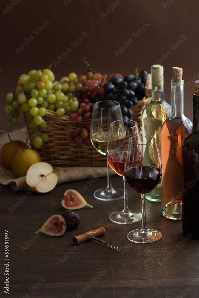 Red, rose and white wine bottles with three wineglasses, grapes in wicker basket and pear apples and figs on brown wood textured table covered with canvas towel