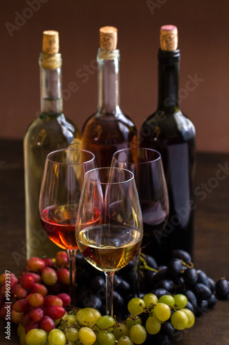 Red, rose and white wine bottles with three wineglasses and grapes on brown wood textured table