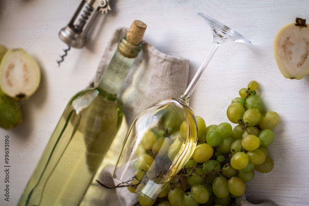 Obraz premium wineglass with white wine with bottle, guava and grapes on white wood textured table covered with canvas towel
