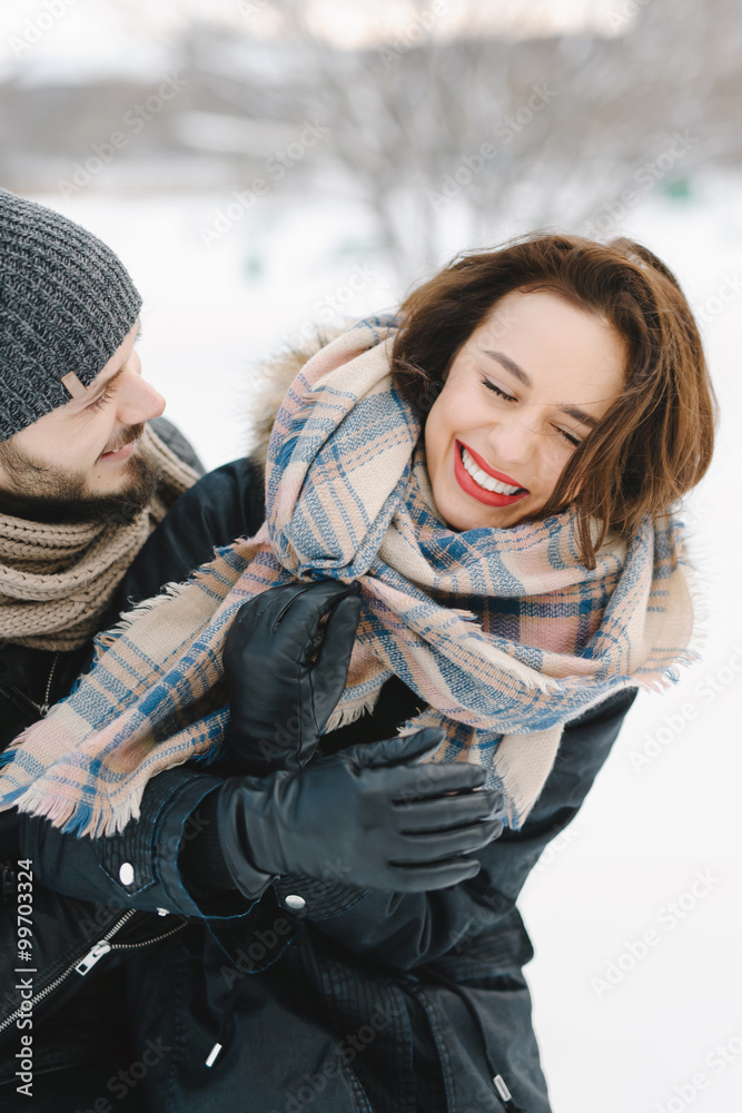 Cute young hipster couple having fun in winter park on a bright day hugging each other and smiling
