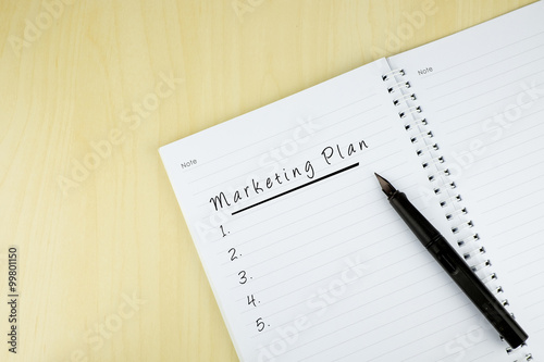 "Marketing Plan" is written on notepad with pen on office table. Top View