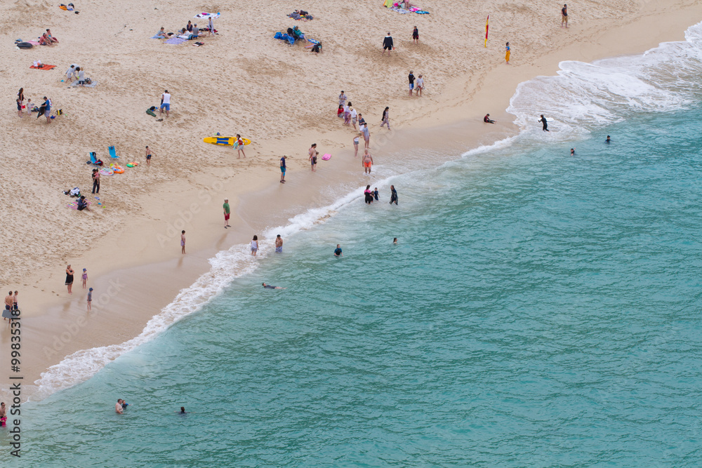 A busy, sandy beach and sea with holidaymakers enjoying themselves. Looking down from above.