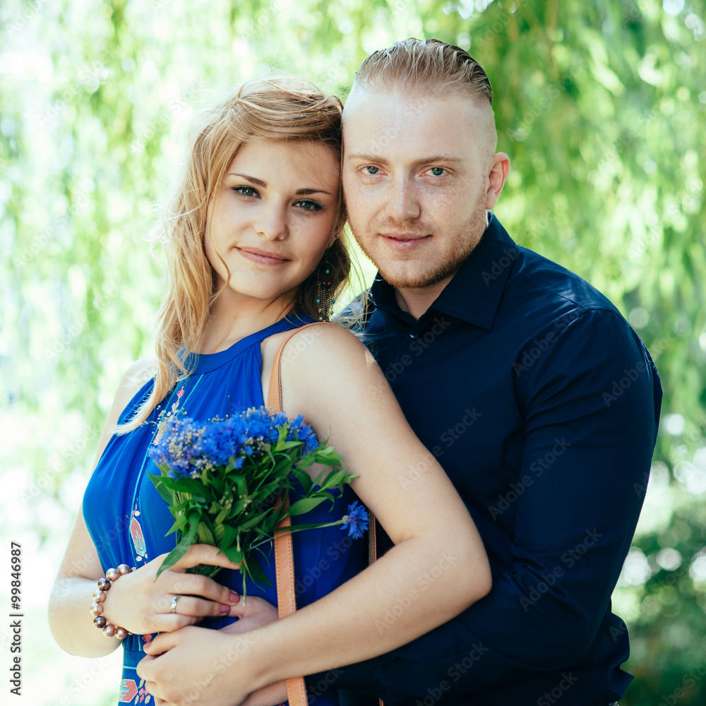 Portrait of blonde young couple against natural scenery. Blue dress with ethnic motives. Stylish man in classic dark outfit. Cornflowers. Portrait of blonde young couple against natural scenery. Blue dress with ethnic motives. Stylish man in classic dark outfit. Cornflowers.
